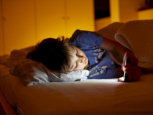 Ein Bub bedient im Bett ein Smartphone - 60 Prozent der Teilnehmer einer Umfrage gaben an, ihr Smartphone mit ins Bett zu nehmen. (Symbolbild)