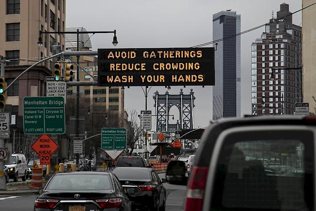 Im Hintergrund eines blinkenden Schilds im Stadtteil Brooklyn ist die Manhattan-Brücke zu sehen. Das Schild fordert Verkehrsteilnehmer auf, Versammlungen und Gedränge zu vermeiden und sich die Hände zu waschen.